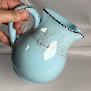 May include: A light blue ceramic pitcher with a curved handle and a spout. The pitcher has a distressed finish with brown accents and a dark brown rim. The pitcher is held by a hand.