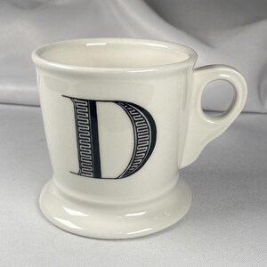 May include: Cream-colored ceramic mug with a black letter "D" on the front. The mug has a handle and a flared base. The letter "D" is in a decorative font.