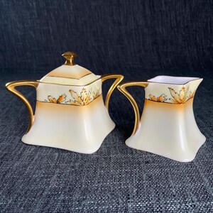 May include: A vintage cream and sugar set. The set includes a lidded sugar bowl and a creamer. Both pieces are white with gold handles and trim. The tops of the pieces have a gradient of orange and yellow with a leaf design.