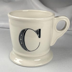 May include: Cream-colored ceramic mug with a black letter "C" on the front. The mug has a rounded base, a handle, and a slightly flared rim. The letter "C" is stylized with decorative details.