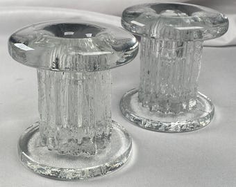 May include: Two clear glass candle holders. Each has a round base, a textured, cylindrical stem, and a flat, circular top with a recessed area for a candle. The glass has a textured, ice-like appearance. The candle holders are displayed on a white surface.