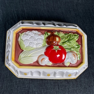 May include: A rectangular ceramic serving dish with a raised relief design of vegetables. The vegetables include a red tomato, a brown onion, cauliflower, lettuce, and mushrooms. The dish has a white and yellow scalloped border.