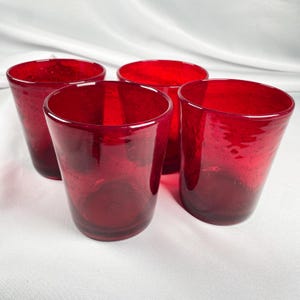May include: A set of four vibrant red drinking glasses featuring a distinct bubble texture throughout the glass material. Each tumbler has a cylindrical shape with a slightly wider rim and a smooth, rounded base, showcasing a deep crimson hue.
