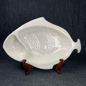 May include: White ceramic fish-shaped serving dish with three compartments. The dish has a textured surface and is supported by a dark brown wooden stand. The dish is designed to resemble a fish.