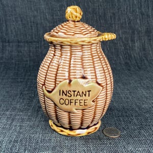 May include: A vintage ceramic instant coffee jar with a lid. The jar is tan and brown with a woven basket design. The words "INSTANT COFFEE" are printed on a leaf-shaped label on the front. The jar has a small handle on the lid.