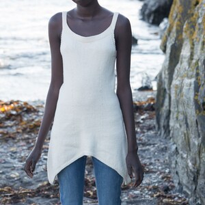 Peut inclure: Une personne portant un débardeur en tricot blanc et un jean bleu se tient sur une plage avec des rochers et de l'eau en arrière-plan.