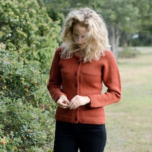 May include: A woman wearing a rust-colored knitted cardigan with a button closure. The cardigan has a textured knit pattern and is worn over a black shirt and black pants.