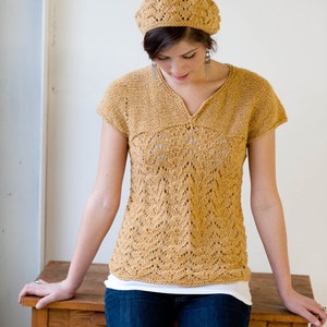 May include: A mustard-colored, short-sleeved knit top and matching beret. The top features a V-neck and a detailed openwork pattern on the front. The beret has a similar openwork design. The person is wearing blue jeans and a white undershirt.