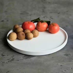 May include: A white, round serving tray displays a selection of fresh fruit. The arrangement features several brown longan fruits, three vibrant red persimmons, and a single red tomato. The tray rests on a grey surface.