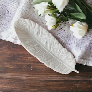 May include: White ceramic feather-shaped dish. The dish is designed to look like a real feather with detailed texture.