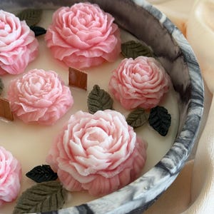 Puede incluir: Una vela decorativa con seis flores de cera rosas en forma de rosa y detalles de hojas verdes. La vela está colocada en una bandeja redonda con diseño de mármol. El fondo es de color claro y neutro.