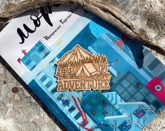 Może przedstawiać: Wycięty z drewna motyw gór z namiotem, drzewami i ogniskiem. Wycięcie jest brązowe i zawiera napis "ADVENTURE" białymi literami.