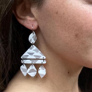 Pode incluir: Brincos geométricos brancos pendentes com forma triangular e múltiplas formas de gota. Os brincos têm um padrão geométrico branco e preto.