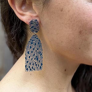 Pode incluir: Um par de brincos estampados em azul e bege. Os brincos têm uma parte superior retangular e uma parte inferior longa e arqueada. O padrão consiste em linhas curvas e semicírculos. Os brincos pendem de um gancho prateado.