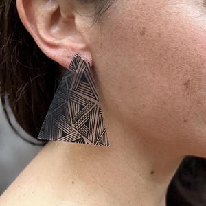 Pode incluir: Um par de brincos em forma de triângulo com um padrão geométrico em preto e branco. Os brincos são feitos de acrílico transparente e têm um design moderno e abstrato.
