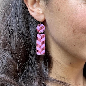 Pode incluir: Brincos geométricos com padrão rosa e vermelho com gancho dourado. Os brincos são feitos de duas peças, uma redonda e uma retangular.