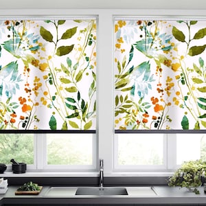 Op de afbeelding: Twee witte raambekleding met een aquarel bloemenpatroon met groene bladeren, blauwe bloemen en gele en oranje bessen. De raambekleding bevindt zich in een keuken met een gootsteen en aanrecht.