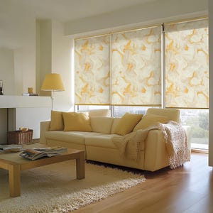 Può includere: Un soggiorno con un grande divano angolare color crema con cuscini e una coperta. Il divano è di fronte a una grande finestra con tre tende a rullo a motivi bianchi e dorati. C'è un tavolino da caffè in legno davanti al divano.