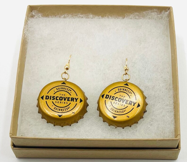 Może przedstawiać: Para złotych kolczyk&oacute;w wykonanych z zakrętek od butelek Guinnessa. Zakrętki mają napis "The Discovery Series" i logo Guinnessa.
