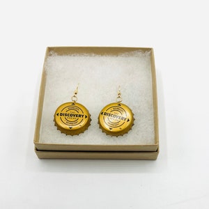 May include: A pair of gold-colored earrings made from repurposed Guinness bottle caps. The bottle caps feature the text "The Discovery Series" and the Guinness logo.