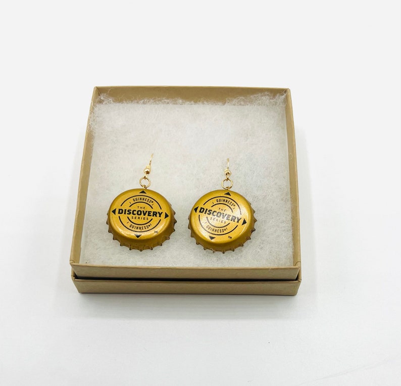 May include: A pair of gold-colored earrings made from repurposed Guinness bottle caps. The bottle caps feature the text "The Discovery Series" and the Guinness logo.
