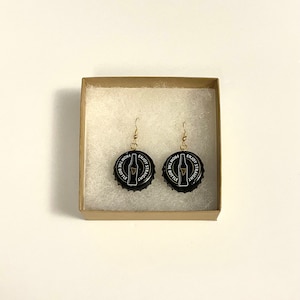May include: A pair of black bottle cap earrings with gold hooks. The bottle caps have a white design that says "Enjoy it Straight from the Bottle" and a picture of a beer bottle.