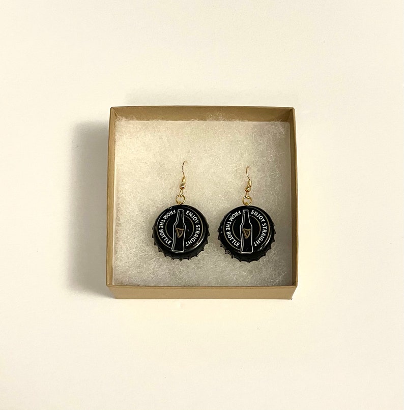 May include: A pair of black bottle cap earrings with gold hooks. The bottle caps have a white design that says "Enjoy it Straight from the Bottle" and a picture of a beer bottle.