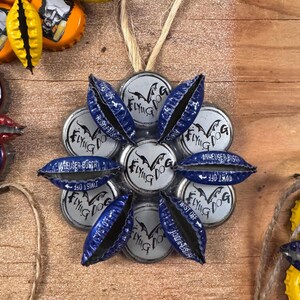 Op de afbeelding: Een zilveren en blauwe ronde ornament gemaakt van flessendoppen. De doppen hebben de woorden "Flying Dog" en "Twist Off" erop gedrukt.