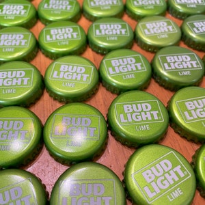 Puede incluir: Primer plano de numerosas tapas de botellas Bud Light Lime verdes. Cada tapa muestra el logotipo de Bud Light en blanco y la palabra "LIME". Las tapas son de color verde metálico y están dispuestas sobre una superficie de madera.