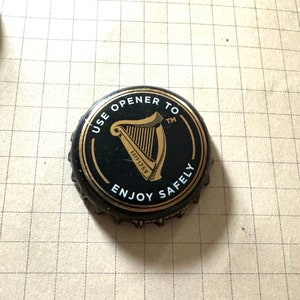 May include: Black bottle cap with gold lettering that reads "Use opener to enjoy safely" and a gold harp logo with "Est 1759" printed below. The text "TM" is printed in the top right corner.