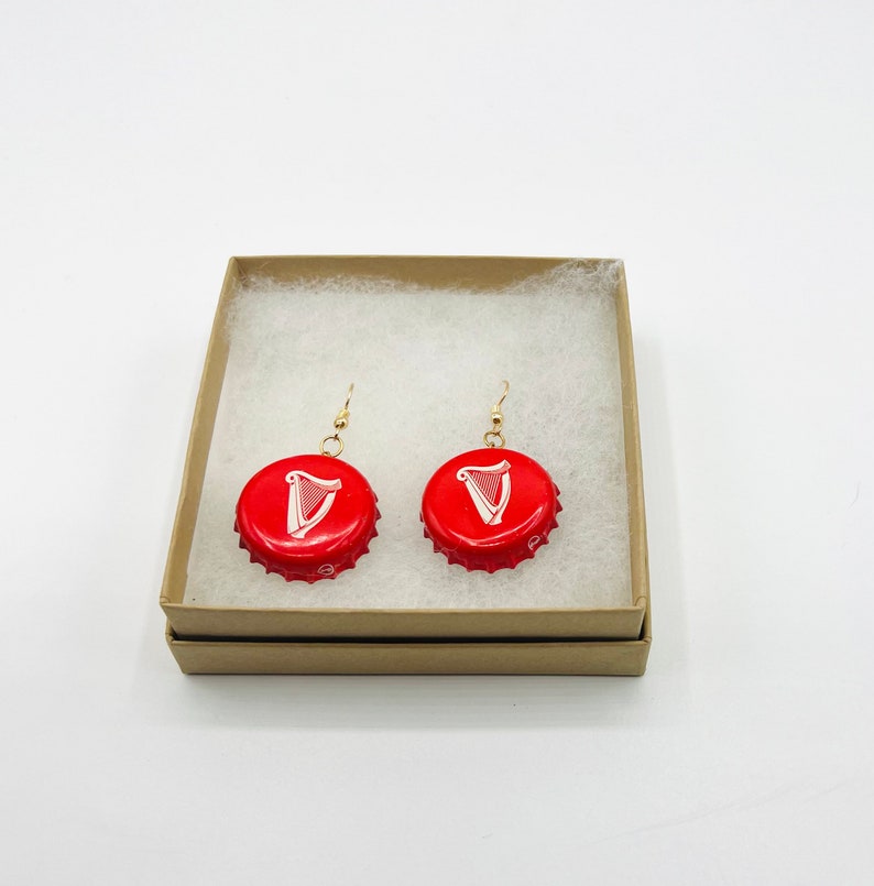 May include: A pair of red bottle cap earrings with a white harp logo. The earrings are displayed in a brown box with white tissue paper.