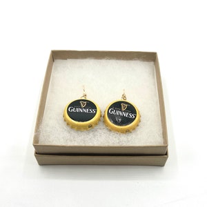 May include: A pair of gold earrings with black Guinness bottle caps. The bottle caps have the Guinness logo and the text "ESTD 1778 GUINNESS" printed on them.