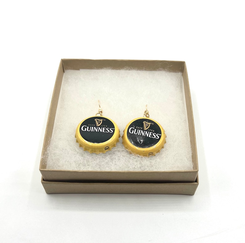 May include: A pair of gold earrings with black Guinness bottle caps. The bottle caps have the Guinness logo and the text "ESTD 1778 GUINNESS" printed on them.