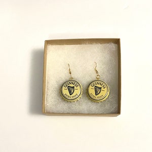 May include: A pair of gold earrings with Guinness bottle caps as the charms. The bottle caps are yellow with black lettering that reads "GUINNESS" and "EXTRA STOUT".
