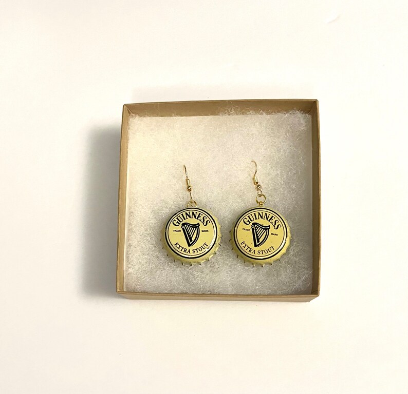 May include: A pair of gold earrings with Guinness bottle caps as the charms. The bottle caps are yellow with black lettering that reads "GUINNESS" and "EXTRA STOUT".