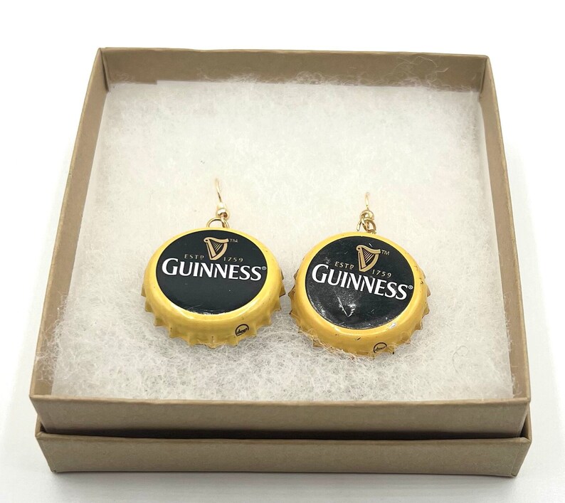 Może przedstawiać: Para złotych kolczyk&oacute;w wykonanych z zakrętek od butelek Guinnessa. Zakrętki są ż&oacute;łte z czarnym napisem "ESTD 1759 GUINNESS TM". Kolczyki znajdują się w małym pudełku z białym wyściełaniem.