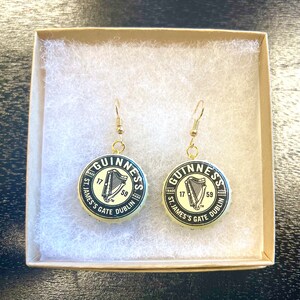 May include: A pair of gold earrings with black and white Guinness bottle cap charms. The charms feature the Guinness logo, a harp, and the text "GUINNESS", "TRADE MARK", "ST. JAMES'S GATE", "DUBLIN", "17", and "59".