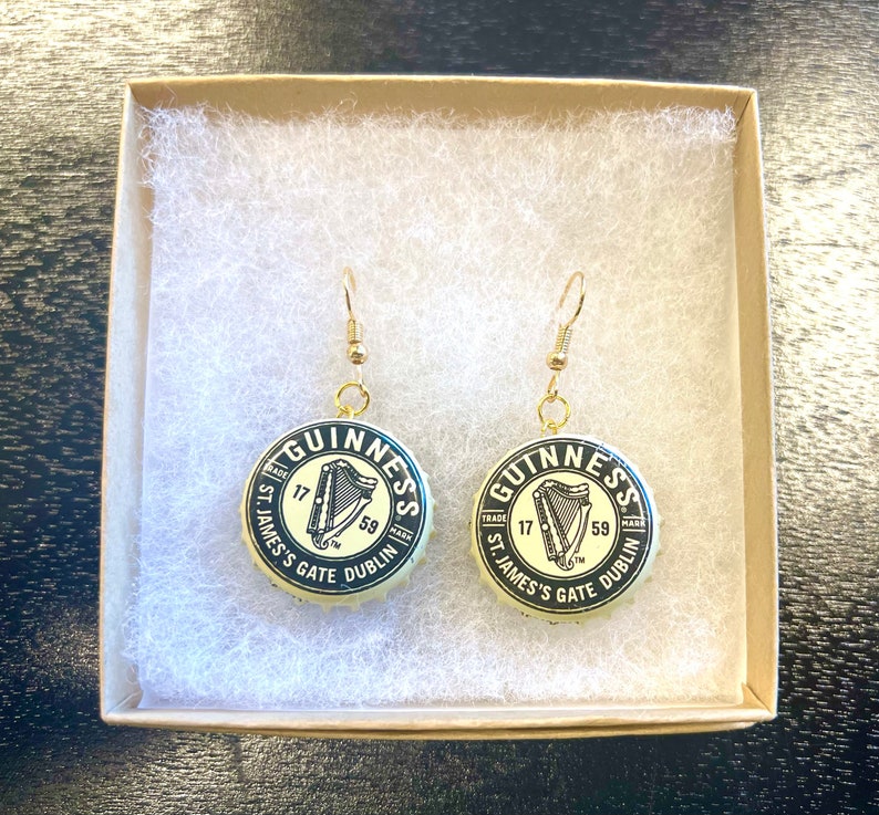 May include: A pair of gold earrings with black and white Guinness bottle cap charms. The charms feature the Guinness logo, a harp, and the text "GUINNESS", "TRADE MARK", "ST. JAMES'S GATE", "DUBLIN", "17", and "59".