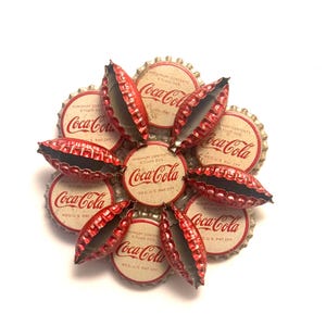 May include: A flower-shaped arrangement made from vintage Coca-Cola bottle caps. The caps are red and silver, with the Coca-Cola logo and text printed on the white center. The arrangement is on a white background.