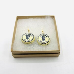 May include: A pair of gold earrings with Guinness Extra Stout bottle caps. The bottle caps are cream colored with a gold rim and feature the Guinness logo and harp.