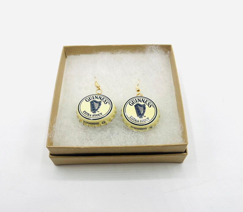 May include: A pair of gold earrings with Guinness Extra Stout bottle caps. The bottle caps are cream colored with a gold rim and feature the Guinness logo and harp.