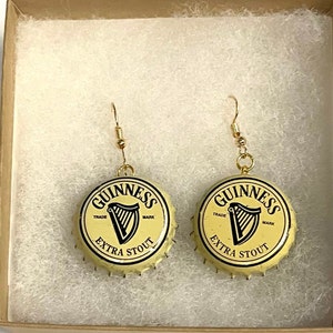 Może przedstawiać: Para złotych kolczyk&oacute;w z korkami od butelek Guinness jako wiszące elementy. Korki od butelek są kremowe z czarnym napisem "GUINNESS TRADE MARK EXTRA STOUT" i czarnym logo harfy.