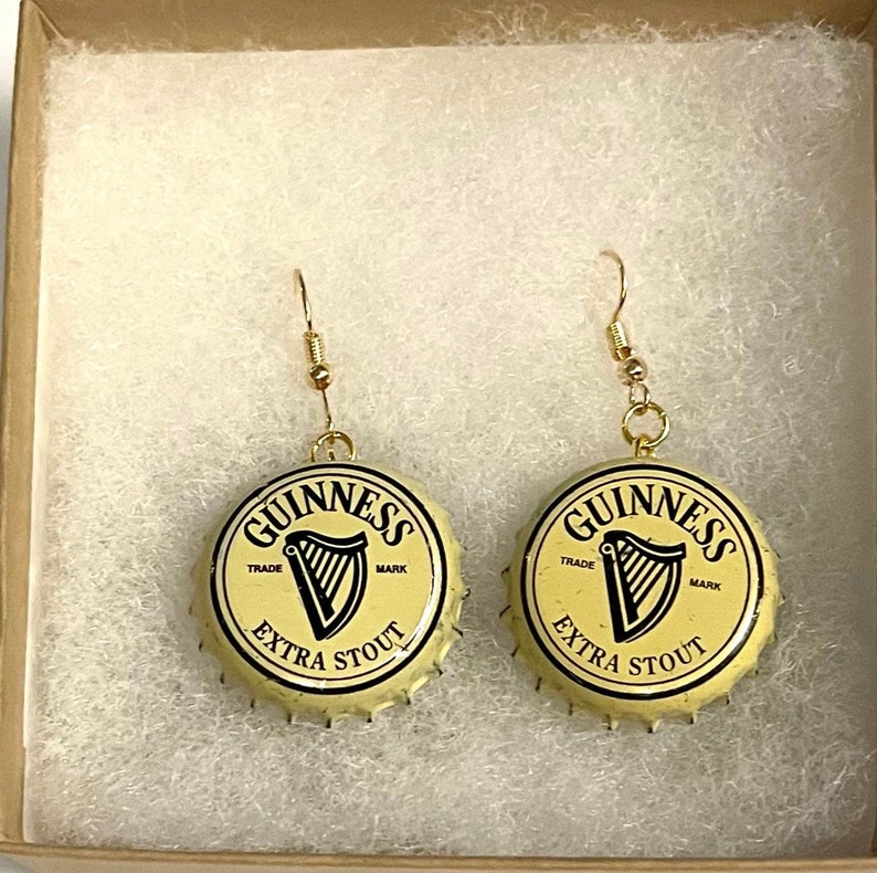 Może przedstawiać: Para złotych kolczyk&oacute;w z korkami od butelek Guinness jako wiszące elementy. Korki od butelek są kremowe z czarnym napisem "GUINNESS TRADE MARK EXTRA STOUT" i czarnym logo harfy.