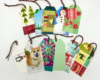 Puede incluir: Un conjunto de diez etiquetas de regalo rectangulares de papel con esquinas recortadas y cordeles rojos y verdes. Los diseños incluyen un muñeco de nieve, un corgi, un pingüino, una chimenea con calcetines y patrones geométricos.