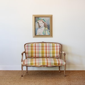 May include: A two-seater settee with a wooden frame and a multi-colored plaid fabric upholstery in shades of pink, yellow, and orange. Above the settee hangs a framed painting depicting a person wearing a floral headpiece and a pearl necklace.