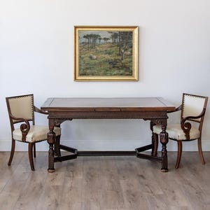 May include: A rectangular dark wood dining table with carved legs and a central stretcher, flanked by two armchairs featuring cream-colored upholstered seats and backs with nailhead trim, and a framed landscape painting hanging on the wall.