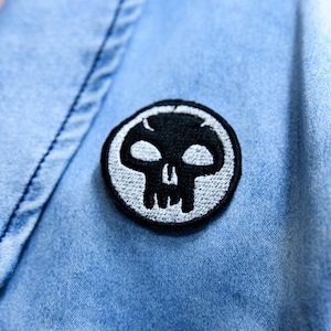 Può includere: Una toppa a forma di teschio in bianco e nero è cucita su una giacca di jeans blu chiaro. La toppa è circolare con un bordo bianco e presenta un design a teschio stilizzato. Il tessuto della giacca ha un aspetto strutturato.