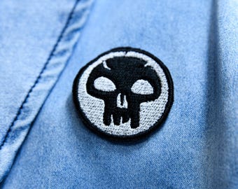 May include: A black and white skull patch is sewn onto a light blue denim jacket. The patch is circular with a white border and features a stylized skull design. The jacket's fabric has a textured appearance.