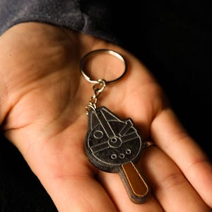 Può includere: Un portachiavi nero e marrone del Millennium Falcon. Il portachiavi è realizzato in un materiale scuro e presenta l'iconico design dell'astronave. È attaccato a un portachiavi argentato.