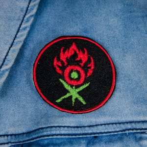 May include: A round, black embroidered patch with a red and green design, affixed to a light blue denim jacket. The design features a stylized flame and floral motif.